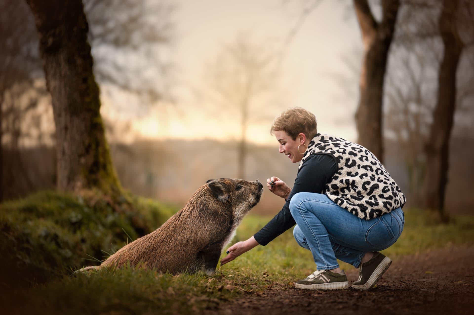 Femme accroupie face à son cochon dans un sous-bois au soleil levant