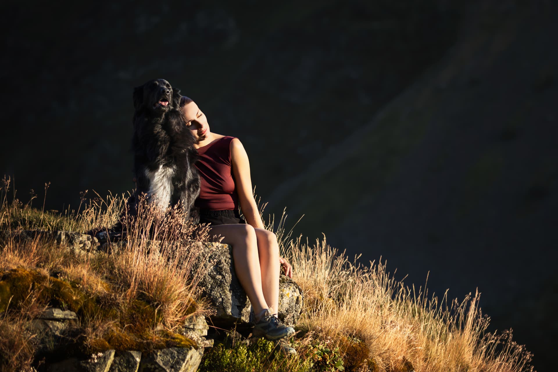 Femme assise sur un rocher au coucher du soleil, la tête appuyée contre son chien