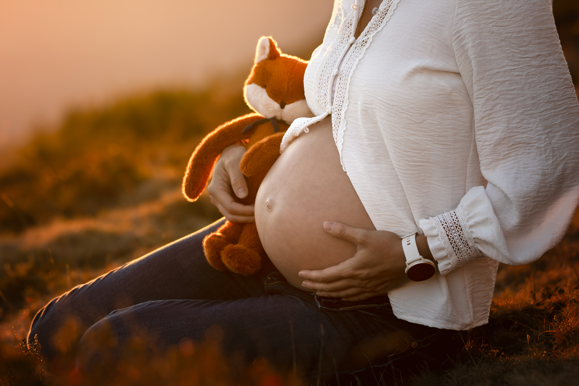 Femme enceinte en blouse blanche serrant son ventre contre un renard en peluche, au coucher du soleil