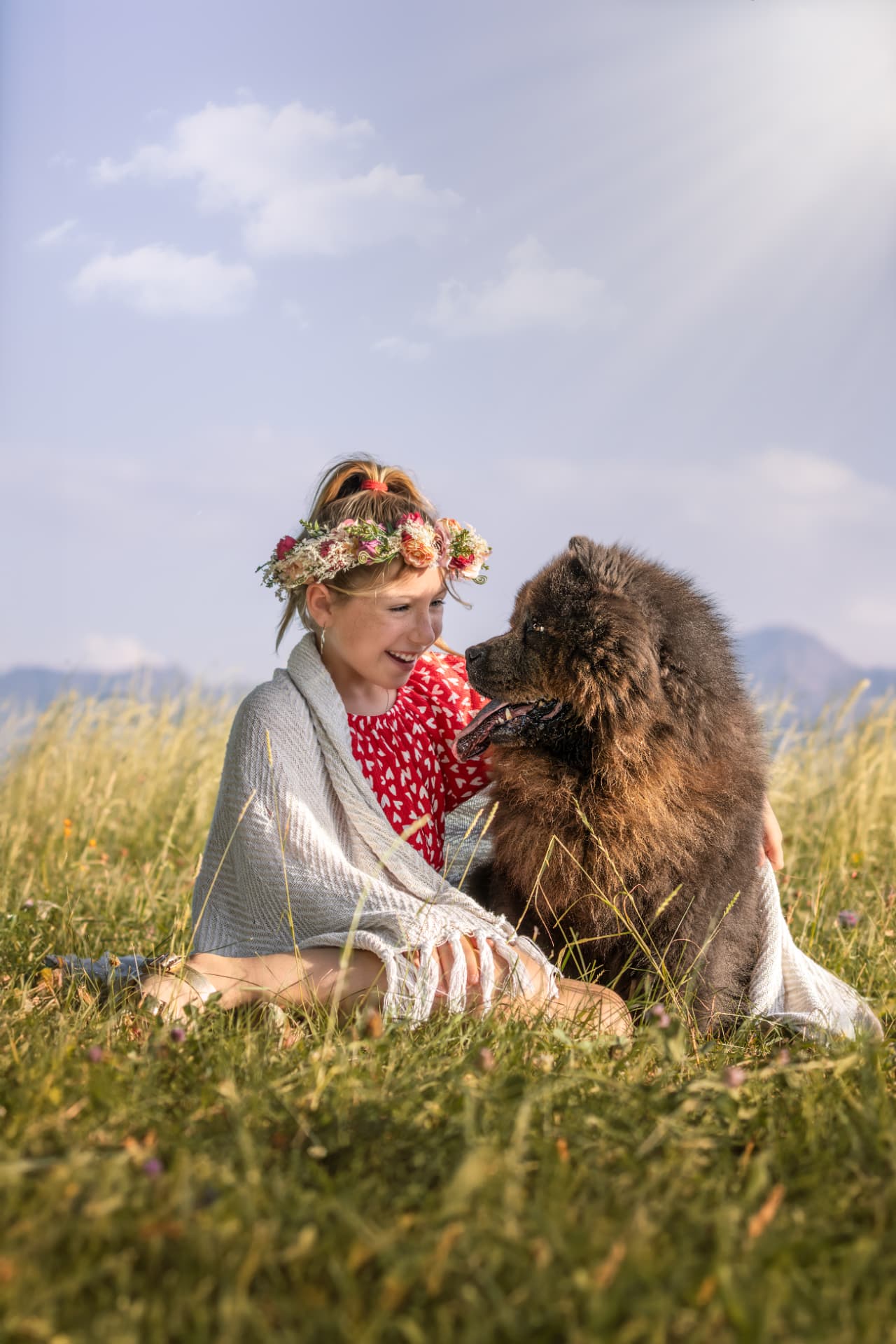 Petite fille couronnée de fleurs face à son chien dans une prairie d’altitude en été